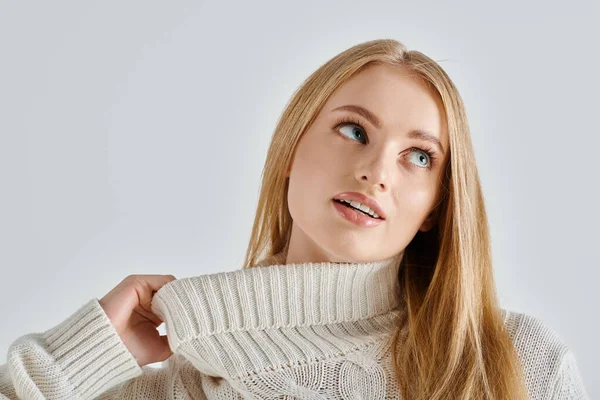 Mujer encantadora y soñadora estiramiento cuello de suéter de punto blanco y mirando hacia otro lado en gris - foto de stock