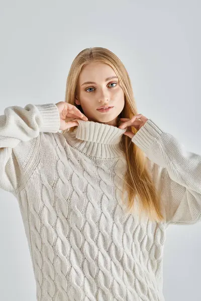 Sensual dreamy woman with blonde hair touching collar of warm sweater and looking at camera on grey — Stock Photo
