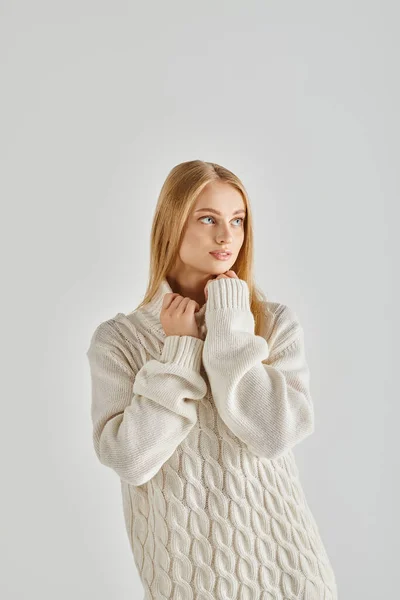 Mujer rubia contemplativa en suéter blanco cálido mirando a la cámara en gris, emociones de invierno - foto de stock