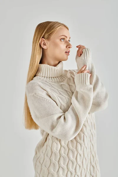 Zierliche und nachdenkliche blonde Frau in weißem Strickpullover, die auf graue, gemütliche Winterbekleidung blickt — Stockfoto
