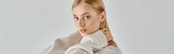 Young blonde woman in soft knitted sweater embracing own neck and looking at camera on grey, banner — Stock Photo