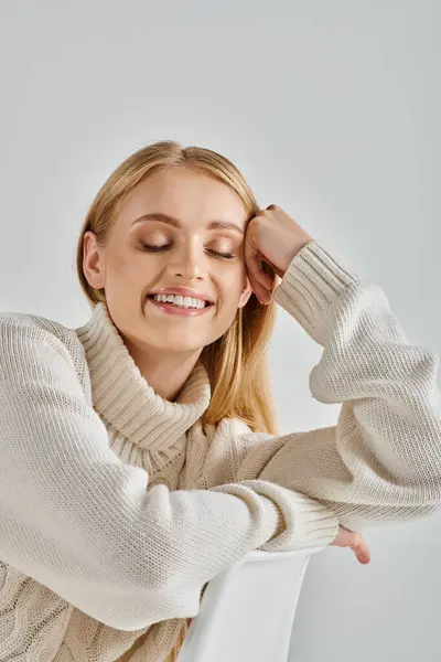 Felice donna bionda in maglione caldo bianco sorridente con gli occhi chiusi su sfondo grigio, gioia invernale — Foto stock