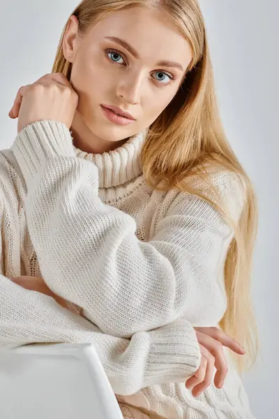 Романтична блондинка в білому затишному светрі сидить і дивиться на камеру на сірому, зимовому шармі — стокове фото