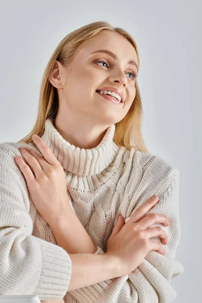 Femme insouciante avec des cheveux blonds assis dans un pull d'hiver confortable et regardant loin sur gris, bonheur — Photo de stock