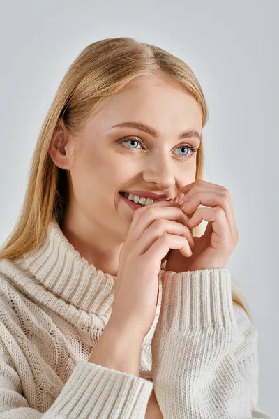 Jeune femme joyeuse en pull blanc chaud et maquillage naturel souriant avec les mains près du visage sur gris — Photo de stock