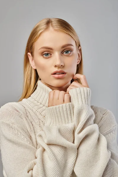 Nachdenkliche Frau mit blonden Haaren und natürlichem Make-up im weißen Winterpullover blickt in die Kamera auf grau — Stockfoto