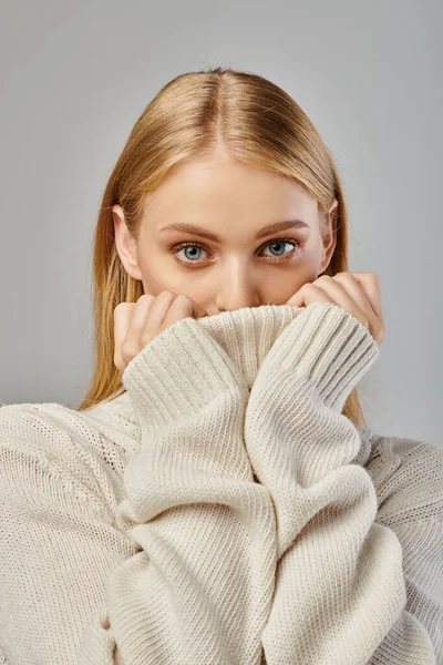 Young blonde woman obscuring face with warm winter sweater on grey backdrop, sophisticated charm — Stock Photo