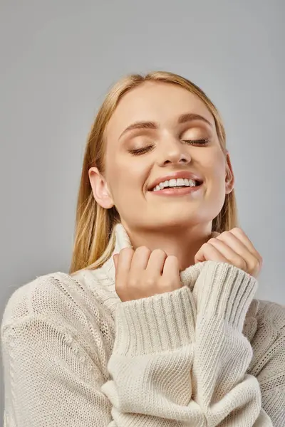 Deliziata donna bionda in caldo maglione a maglia sorridente con gli occhi chiusi sul grigio, moda invernale — Foto stock