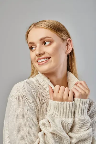Mujer rubia alegre en suéter de punto blanco sonriendo y mirando hacia otro lado en gris, felicidad de invierno - foto de stock