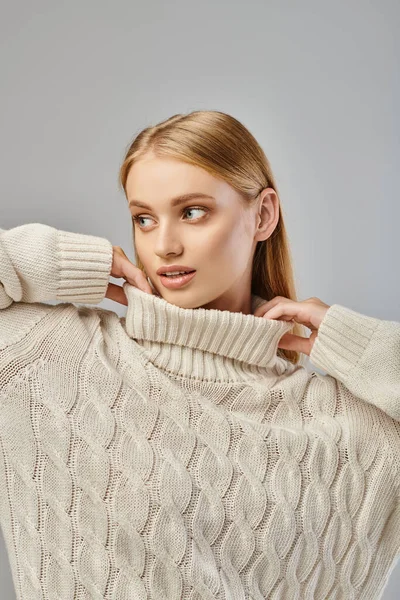 Dreamy blonde woman with natural makeup touching collar of white sweater and looking away on grey — Stock Photo