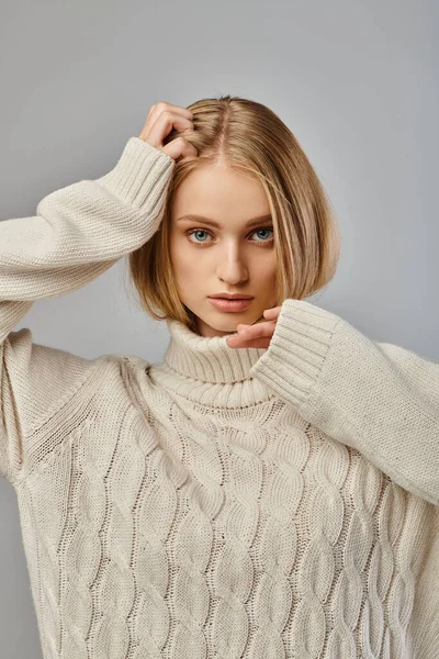 Молодая женщина с светлыми волосами и выразительным взглядом позирует в белом трикотажном свитере на сером фоне — стоковое фото