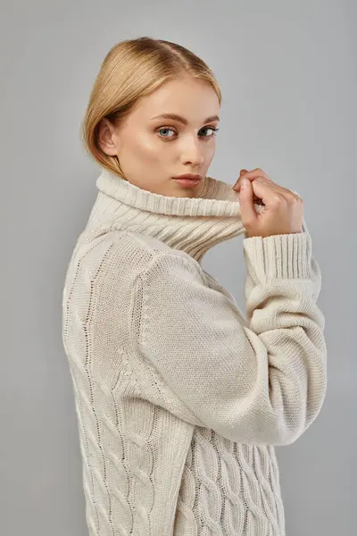 Young blonde woman in warm knitted sweater with high collar looking at camera on grey, winter trend — Stock Photo