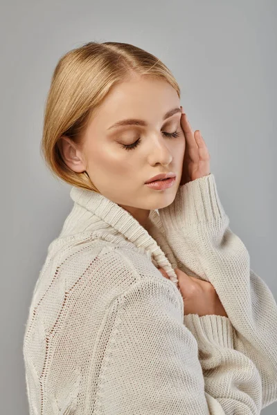 Jeune femme blonde avec maquillage naturel et les yeux fermés posant en pull tricoté blanc sur gris — Photo de stock