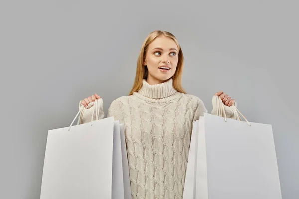 Cheerful blonde woman in warm knitted sweater holding white shopping bags and looking away on grey — Stock Photo