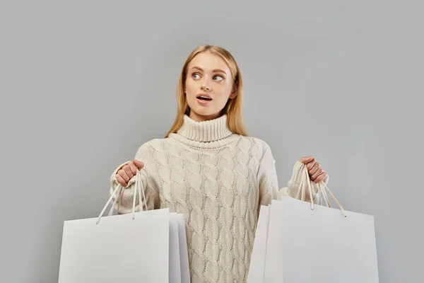Mulher loira espantada em suéter quente branco segurando sacos de compras brancos em cinza, vendas de inverno — Fotografia de Stock