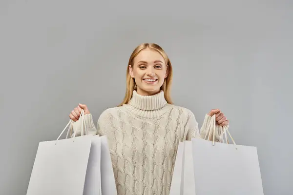 Pleased blonde woman in cozy knitted sweater holding white shopping bags on grey, winter purchases — Stock Photo