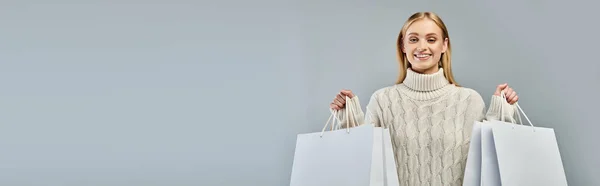 Mulher loira alegre em suéter de malha quente segurando sacos de compras brancos em cinza, bandeira horizontal — Fotografia de Stock
