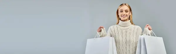 Mulher loira alegre em suéter de malha quente segurando sacos de compras brancos em cinza, bandeira horizontal — Fotografia de Stock