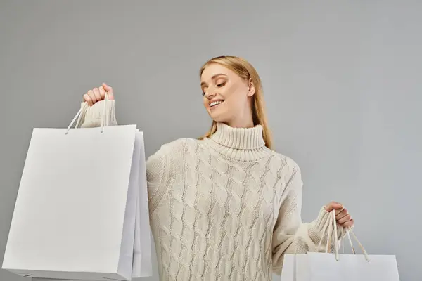Mujer rubia alegre en suéter de punto de invierno posando con bolsas de compras blancas en gris, las ventas de invierno - foto de stock