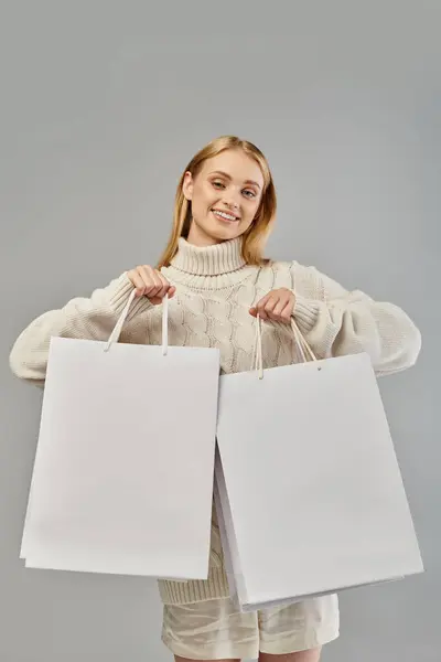 Joyeuse femme blonde en pull tricoté blanc montrant des sacs à provisions sur gris, ventes saisonnières — Photo de stock