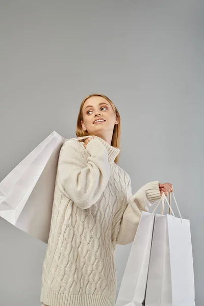 Smiling blonde woman in soft knitted sweater holding white shopping bags and looking away on grey — Stock Photo