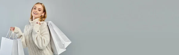 Happy blonde woman in warm knitted sweater posing with white shopping bags on grey, banner — Stock Photo
