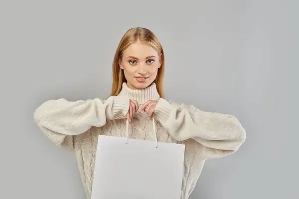Pleased blonde woman in white knitted sweater holding shopping bag and looking at camera on grey — Stock Photo