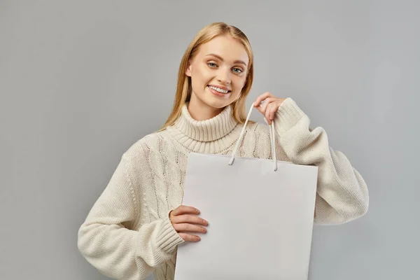 Glad blonde woman in warm knitted sweater holding white shopping bag on grey, winter sales — Stock Photo