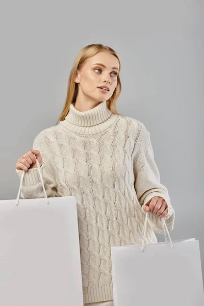 Mesmerizing blonde woman in cozy knitted sweater with white shopping bags looking away on grey — Stock Photo