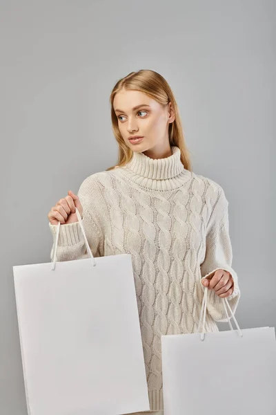 Mujer rubia joven en suéter de punto blanco sosteniendo bolsas de compras y mirando hacia otro lado en gris - foto de stock