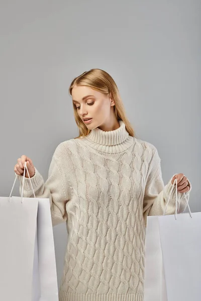Jolie femme blonde en pull élégant chaud avec des sacs à provisions blancs sur gris, achats saisonniers — Photo de stock