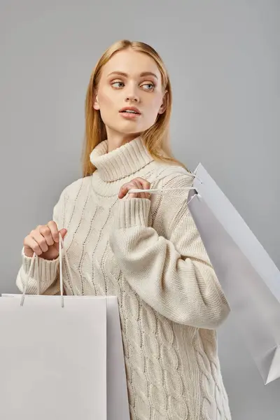 Mulher loira elegante em camisola de malha branca com sacos de compras olhando para longe em cinza, vendas de inverno — Fotografia de Stock