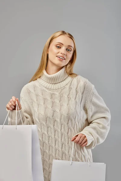 Blonde Frau im Strickwinterpullover mit Einkaufstaschen blickt vor grauem Hintergrund in die Kamera — Stockfoto