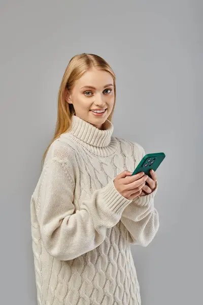 Mujer joven feliz en suéter de punto blanco mensajería en el teléfono inteligente en gris, emociones de invierno - foto de stock