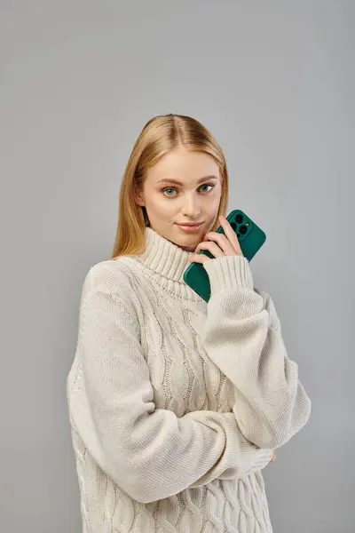 Positive blonde Frau in warmem Strickpullover mit Smartphone und Blick in die Kamera auf grau — Stockfoto