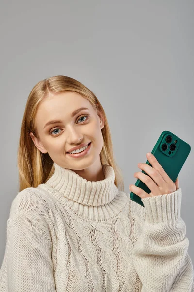 Giovane e allegra donna bionda in maglione invernale che tiene lo smartphone e guarda la fotocamera sul grigio — Foto stock