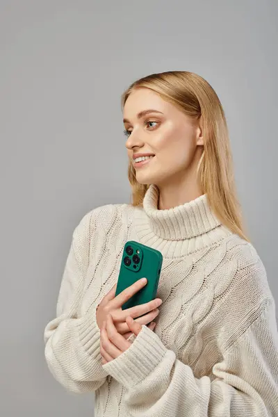 Smiling and dreamy woman in delicate knitted sweater holding  smartphone and looking away on grey — Stock Photo