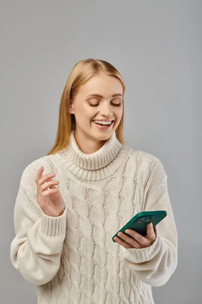 Cheerful blonde woman in warm knitted sweater smiling during video call on smartphone on grey — Stock Photo