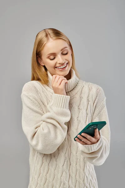 Femme blonde souriante en pull tricoté ayant appel vidéo sur smartphone sur gris, émotions hivernales — Photo de stock