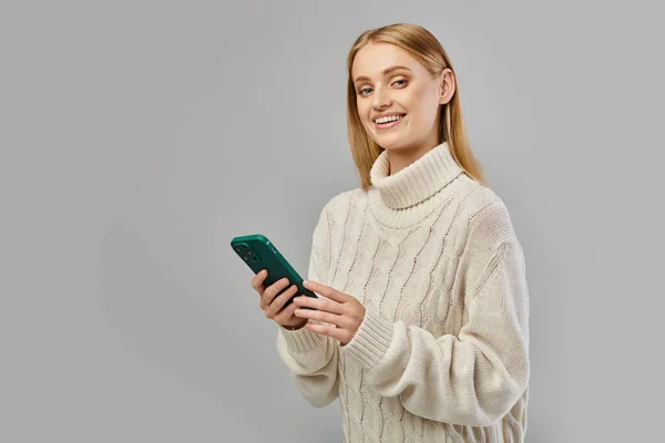 Joyful blonde woman in white winter sweater holding smartphone and smiling at camera on grey — Stock Photo