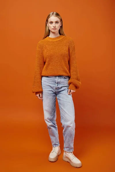 Full length of stylish blonde woman in knitted sweater and blue jeans on orange backdrop, winter — Stock Photo