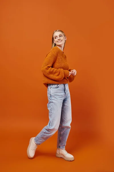 Mujer rubia alegre en suéter de invierno brillante y pantalones vaqueros azules mirando hacia otro lado en el fondo naranja - foto de stock
