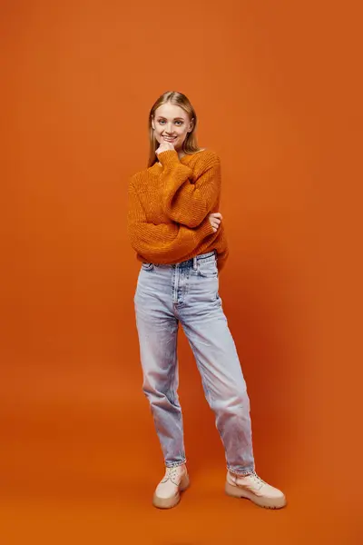 Joyful blonde woman in orange winter sweater and blue jeans smiling at camera on bright backdrop — Stock Photo