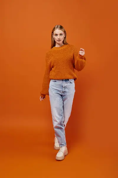 Femme blonde tendance en jeans bleus et pull d'hiver orange marchant sur fond lumineux, pleine longueur — Photo de stock