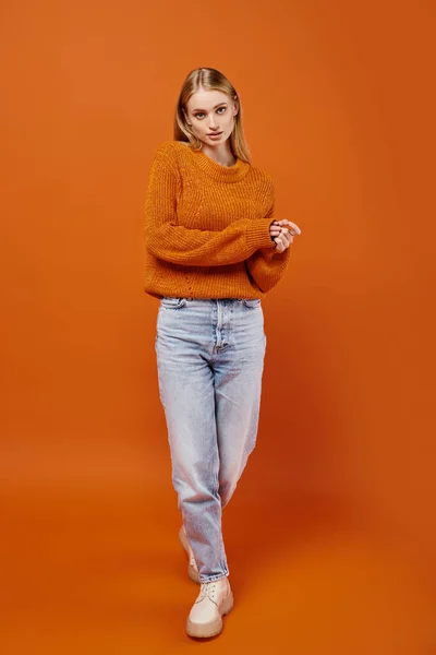 Full length of blonde woman in blue jeans and knitted sweater on orange backdrop, winter fashion — Stock Photo