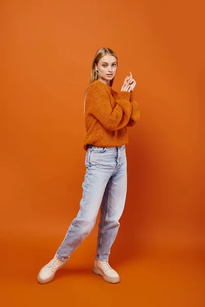 Full length of charming blonde woman in blue jeans and warm sweater on orange backdrop, winter style — Stock Photo