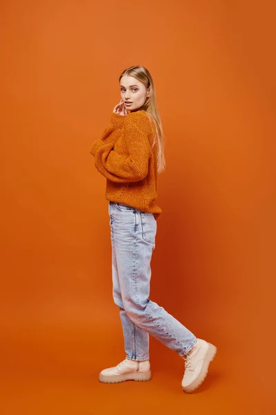 Sensual blonde woman in blue jeans and cozy winter sweater looking at camera on orange backdrop — Stock Photo