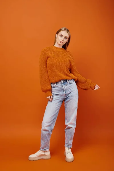 Femme blonde à la mode en jeans et pull en tricot orange posant sur fond lumineux, vibrations hivernales — Photo de stock