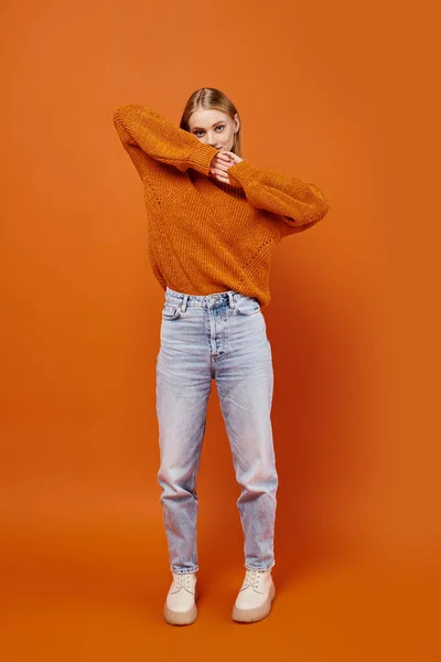 Mujer rubia con estilo en jeans y suéter de punto naranja mirando a la cámara en el fondo brillante - foto de stock