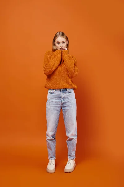 Joyful blonde woman in jeans and warm knitted sweater smiling and looking away on orange backdrop — Stock Photo